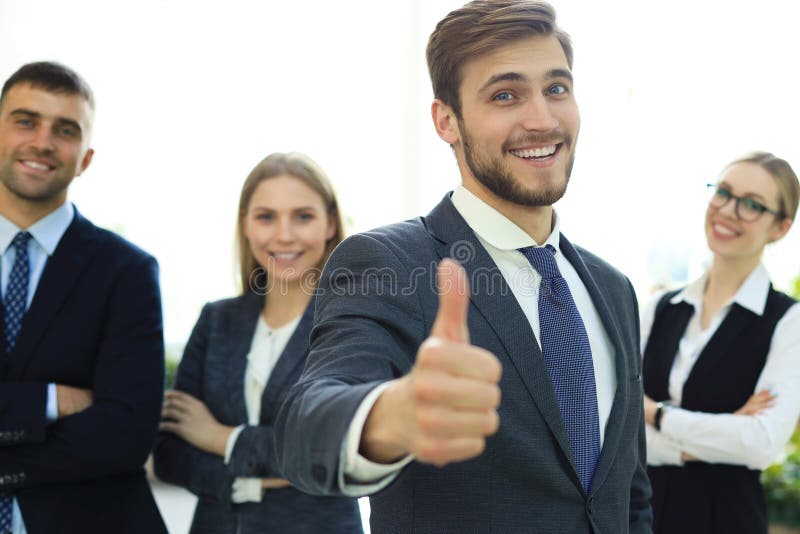 Hombre de negocios feliz que muestra su pulgar para arriba y que sonr?e mientras que sus colegas que se colocan en el fondo fotos de archivo Hombre de negocios feliz que muestra su pulgar para arriba y que sonr?e mientras que sus colegas que se colocan en el fondo fotos de archivo