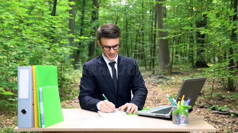 Hombre de negocios inspirado que escribe sus ideas en el cuaderno, sentándose en el escritorio en bosque fotografía de archivo libre de regalías Hombre de negocios inspirado que escribe sus ideas en el cuaderno, sentándose en el escritorio en bosque fotografía de archivo libre de regalías