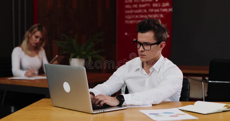 Hombre de negocios joven en vidrios con el ordenador portátil y el papel que trabajan en oficina Negocio, gente, papeleo y