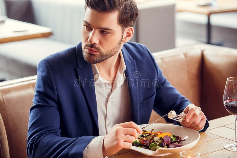 Hombre de negocios joven que se sienta en el restaurante que come el vino de consumición de la ensalada que parece a un lado pens fotografía de archivo Hombre de negocios joven que se sienta en el restaurante que come el vino de consumición de la ensalada que parece a un lado pens fotografía de archivo