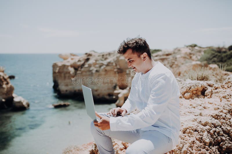 Hombre de negocios joven que usa el ordenador portátil en la playa en fondo de la vista al mar foto de archivo Hombre de negocios joven que usa el ordenador portátil en la playa en fondo de la vista al mar foto de archivo