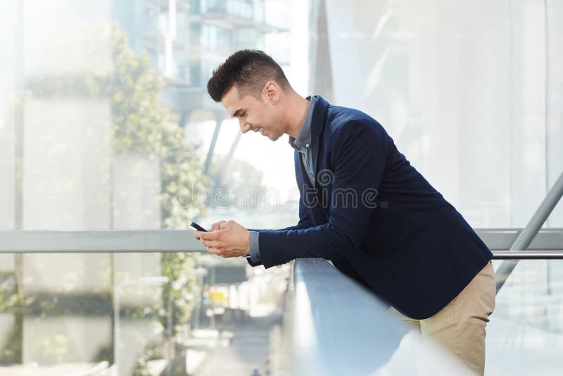 Hombre de negocios joven sonriente que se coloca con el teléfono elegante imágenes de archivo libres de regalías Hombre de negocios joven sonriente que se coloca con el teléfono elegante imágenes de archivo libres de regalías