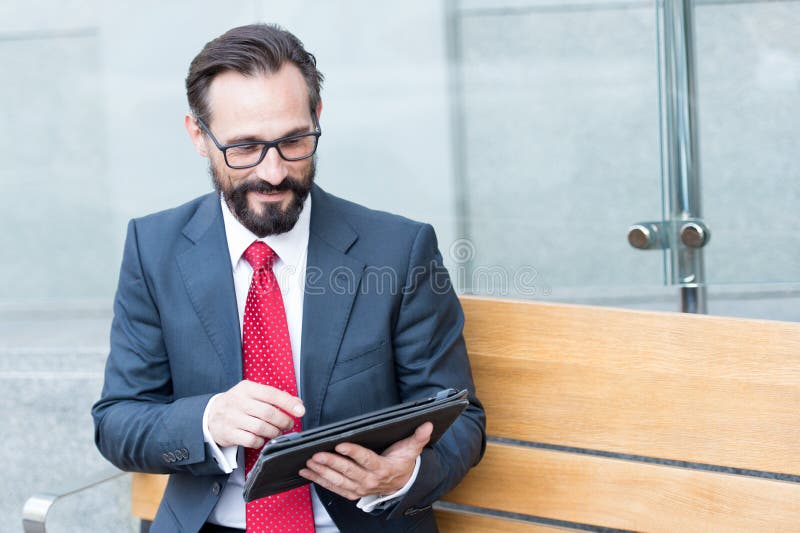 Hombre de negocios positivo elegante que usa una tableta mientras que se sienta en banco imagenes de archivo Hombre de negocios positivo elegante que usa una tableta mientras que se sienta en banco imagenes de archivo