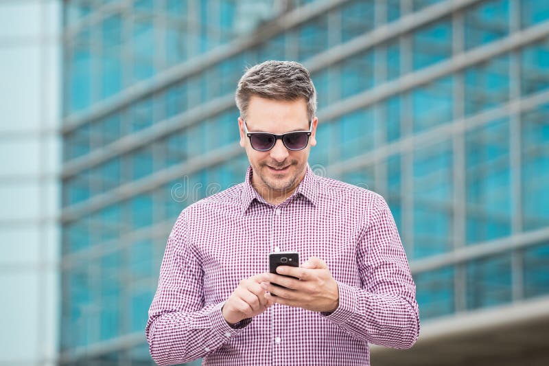 Hombre de negocios que sostiene el teléfono móvil con el edificio de oficinas en el fondo imágenes de archivo libres de regalías Hombre de negocios que sostiene el teléfono móvil con el edificio de oficinas en el fondo imágenes de archivo libres de regalías