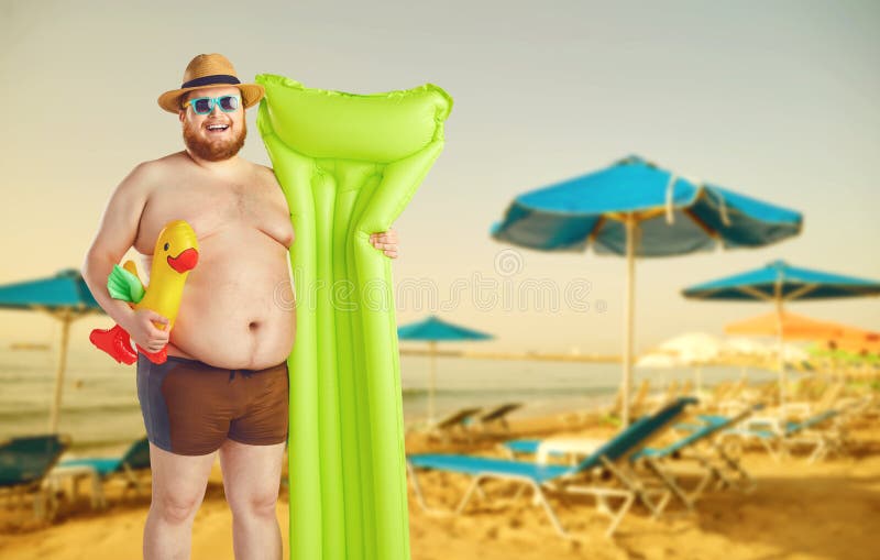 Hombre divertido gordo en troncos de natación con un colchón inflable en un fondo gris imagenes de archivo Hombre divertido gordo en troncos de natación con un colchón inflable en un fondo gris imagenes de archivo