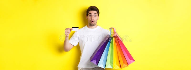 Hombre emocionado haciendo compras en el viernes negro sosteniendo bolsas de papel y tarjeta de crédito parados en el fondo amaril foto de archivo