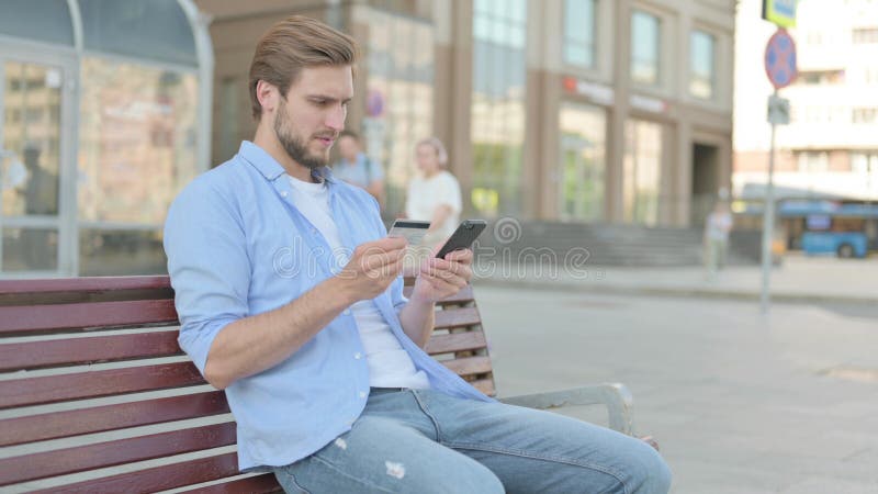 Hombre emocionado haciendo compras en línea vía smartphone mientras estaba sentado al aire libre en el banco imagen de archivo