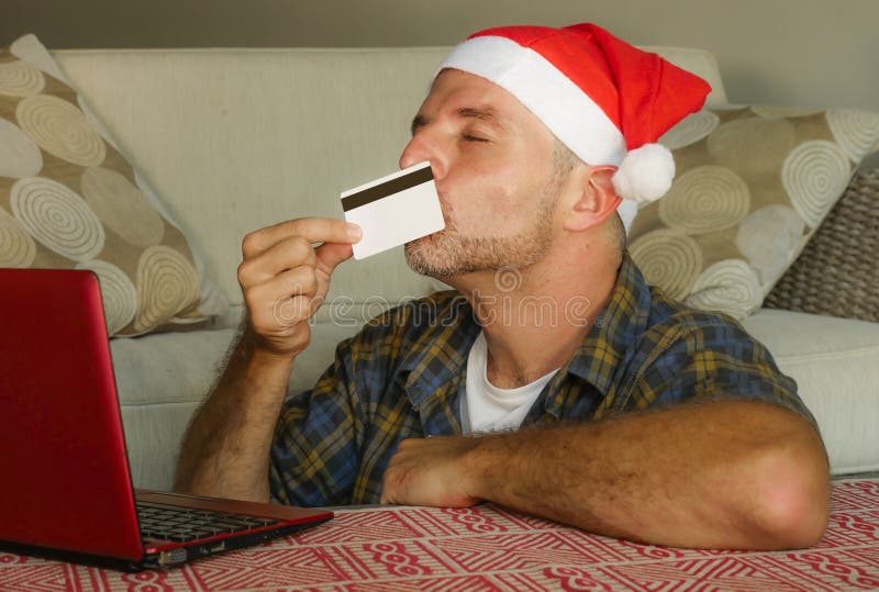 Hombre feliz hermoso y atractivo joven en compras en línea del sombrero de Papá Noel en casa en Internet con el ordenador portáti imagen de archivo libre de regalías Hombre feliz hermoso y atractivo joven en compras en línea del sombrero de Papá Noel en casa en Internet con el ordenador portáti imagen de archivo libre de regalías