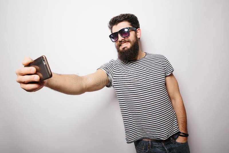 Hombre feliz sonriente del inconformista en vidrios de sol con la barba que toma el selfie con el teléfono móvil imagen de archivo Hombre feliz sonriente del inconformista en vidrios de sol con la barba que toma el selfie con el teléfono móvil imagen de archivo