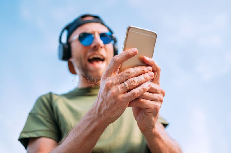 Hombre fresco en gorra de béisbol, auriculares inalámbricos y gafas de sol azules hojeando en su dispositivo del smartphone de la foto de archivo Hombre fresco en gorra de béisbol, auriculares inalámbricos y gafas de sol azules hojeando en su dispositivo del smartphone de la foto de archivo