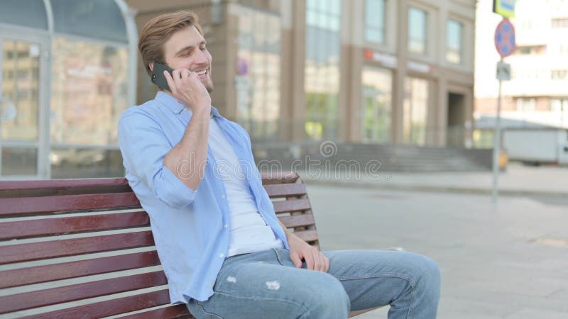 Hombre hablando por teléfono mientras está sentado al aire libre en el banco fotografía de archivo libre de regalías