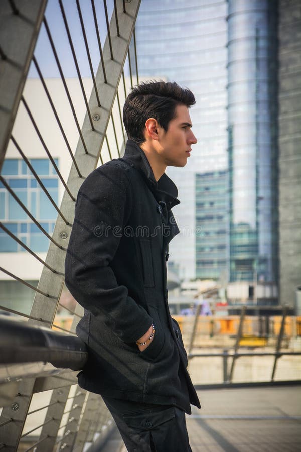 Hombre hermoso joven elegante en la capa negra que se coloca en calle del centro de ciudad imagen de archivo Hombre hermoso joven elegante en la capa negra que se coloca en calle del centro de ciudad imagen de archivo