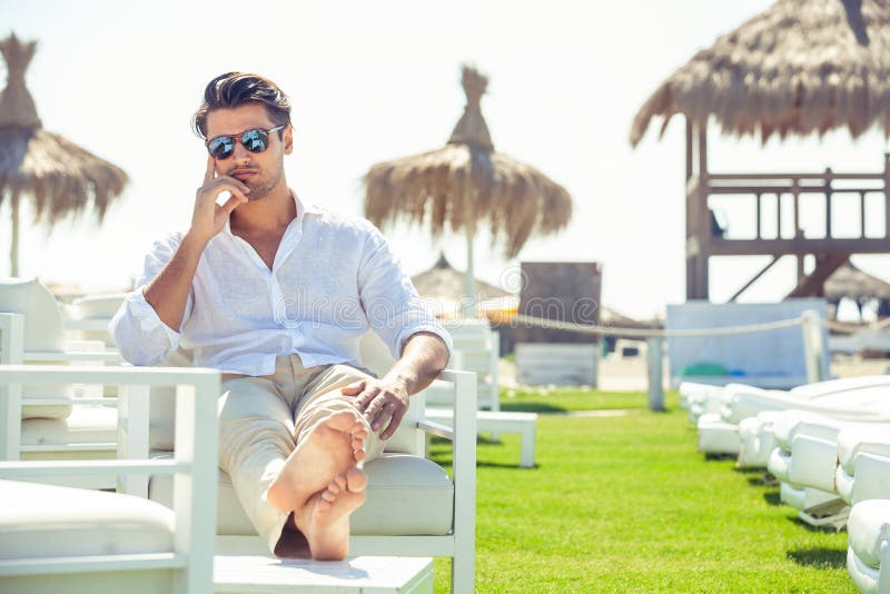Hombre hermoso relajado que se sienta en las sillas blancas durante verano imagen de archivo libre de regalías Hombre hermoso relajado que se sienta en las sillas blancas durante verano imagen de archivo libre de regalías