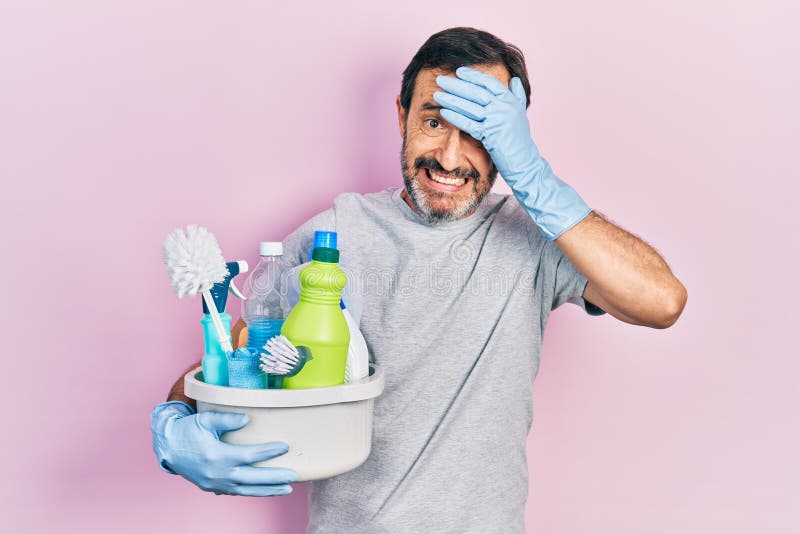 Hombre hispano de mediana edad sosteniendo productos de limpieza estresado y frustrado con la mano en la cabeza sorprendido y rost fotos de archivo libres de regalías