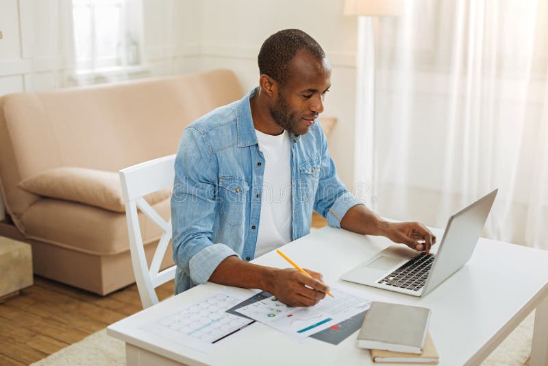 Working. Handsome smiling young afro-american businessman working on his laptop and holding a pencil while sitting at the table. Working. Handsome smiling young afro-american businessman working on his laptop and holding a pencil while sitting at the table