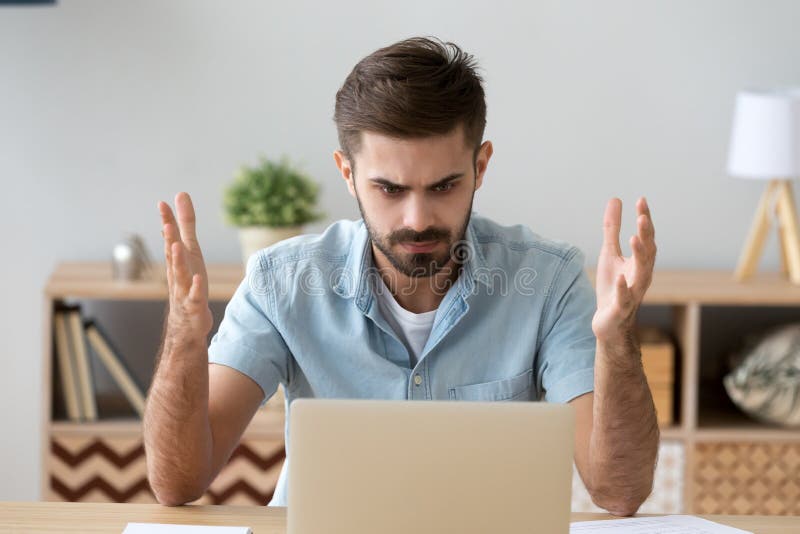 Hombre irritado que mira la pantalla del ordenador portátil que se sienta en el escritorio imagenes de archivo