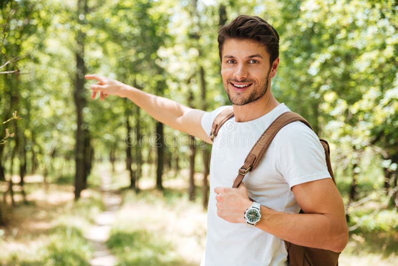 Hombre joven alegre con la mochila que señala lejos en bosque fotografía de archivo Hombre joven alegre con la mochila que señala lejos en bosque fotografía de archivo