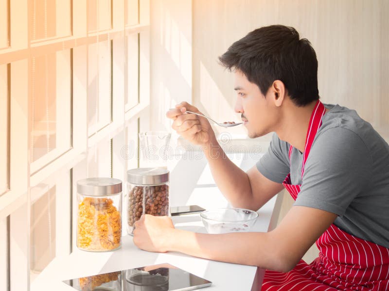 Hombre joven asiático que se sienta al lado del vidrio de la ventana y que come cereal con leche en la tabla para el desayuno en foto de archivo libre de regalías Hombre joven asiático que se sienta al lado del vidrio de la ventana y que come cereal con leche en la tabla para el desayuno en foto de archivo libre de regalías
