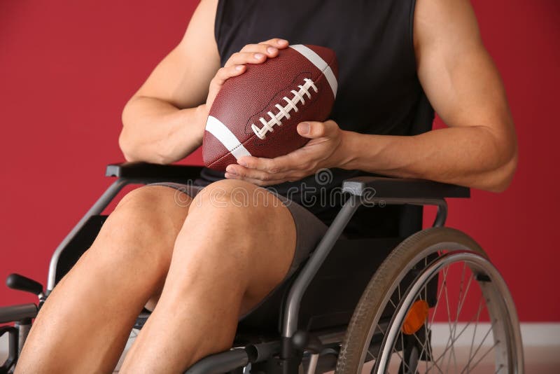 Hombre joven con la bola de rugbi que se sienta en silla de ruedas contra la pared del color fotografía de archivo libre de regalías Hombre joven con la bola de rugbi que se sienta en silla de ruedas contra la pared del color fotografía de archivo libre de regalías