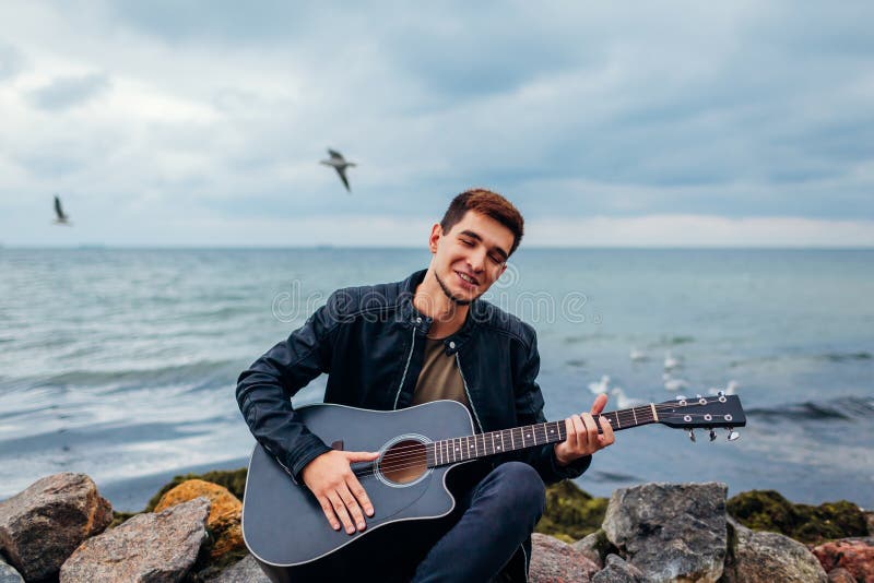 Hombre joven con la guitarra acústica que juega y que canta en la playa rodeada con las rocas en día lluvioso imagen de archivo Hombre joven con la guitarra acústica que juega y que canta en la playa rodeada con las rocas en día lluvioso imagen de archivo