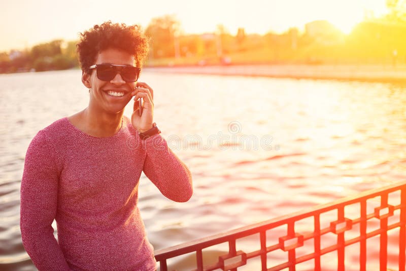 Hombre joven del africano negro con el teléfono celular sobre luz del sol imagen de archivo libre de regalías Hombre joven del africano negro con el teléfono celular sobre luz del sol imagen de archivo libre de regalías