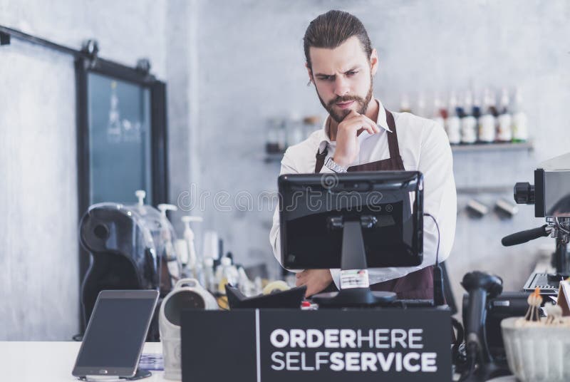 Hombre joven del barista joven confiado que busca un Digital Equipment para pagar el negocio La máquina tiene un problema, ningun fotografía de archivo libre de regalías Hombre joven del barista joven confiado que busca un Digital Equipment para pagar el negocio La máquina tiene un problema, ningun fotografía de archivo libre de regalías