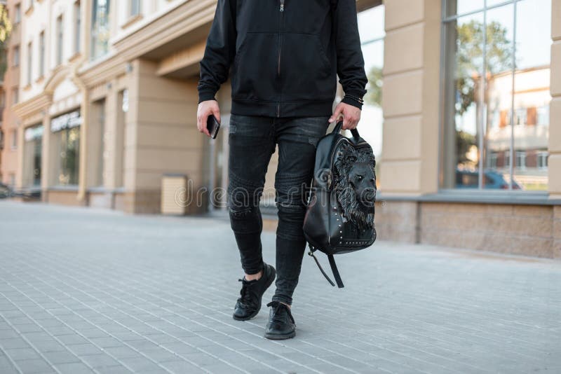 Hombre Joven Elegante En Una Camiseta Negra De Moda Con Una Mochila Negra De Cuero Con Un Modelo Del León En Paseos De Los Vaquer Imagen de archivo - Imagen elegante,