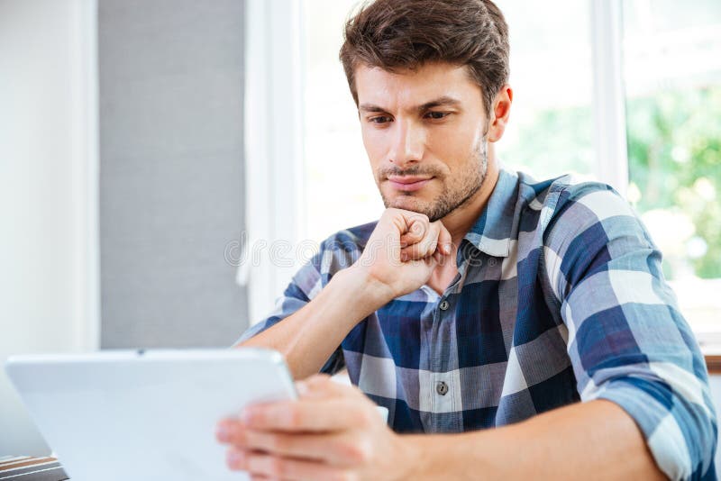 Hombre joven pensativo que piensa y que usa la tableta en casa foto de archivo libre de regalías Hombre joven pensativo que piensa y que usa la tableta en casa foto de archivo libre de regalías