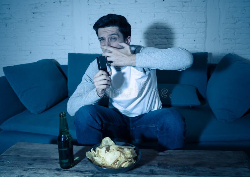 Hombre joven que considera la sentada asustada en el sofá que ve la TV la noche En reacciones y emociones humanas fotografía de archivo libre de regalías Hombre joven que considera la sentada asustada en el sofá que ve la TV la noche En reacciones y emociones humanas fotografía de archivo libre de regalías