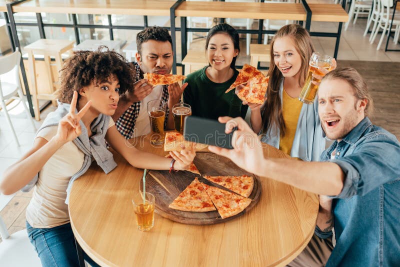 Hombre joven que toma el selfie con los amigos multiétnicos que comen pizza imagenes de archivo Hombre joven que toma el selfie con los amigos multiétnicos que comen pizza imagenes de archivo