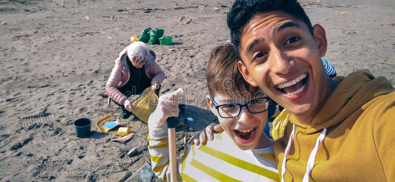 Hombre joven y muchacho que toman el selfie mientras que grupo de voluntarios que limpian la playa imagen de archivo Hombre joven y muchacho que toman el selfie mientras que grupo de voluntarios que limpian la playa imagen de archivo