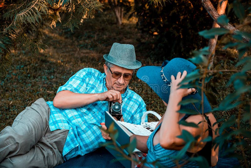 Hombre mayor que fotografía a su esposa mientras que lee un libro en el parque imagen de archivo Hombre mayor que fotografía a su esposa mientras que lee un libro en el parque imagen de archivo