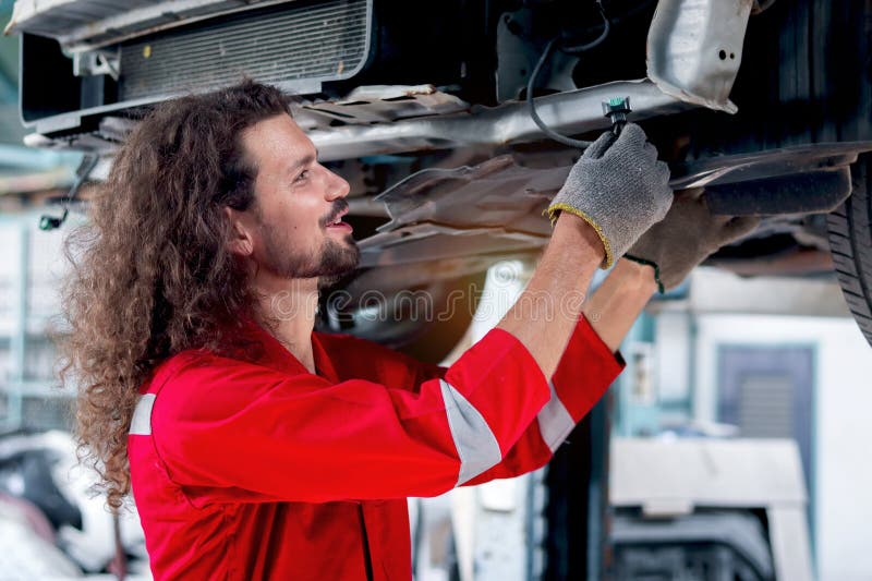 Hombre mecánico de pelo largo con uniforme rojo trabajo con vehículo levantado inspeccionar el sistema de motor de coche de la rue fotografía de archivo libre de regalías