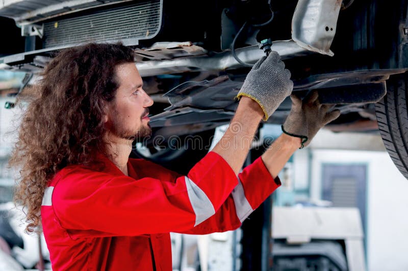Hombre mecánico de pelo largo con uniforme rojo trabajo con vehículo levantado inspeccionar el sistema de motor de coche de la rue imagenes de archivo