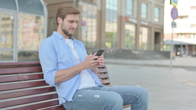 Hombre navegando por Internet en un smartphone mientras estaba sentado al aire libre en el banco fotos de archivo libres de regalías