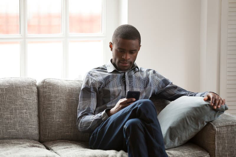 Hombre negro con el teléfono que se sienta en el sofá en casa imagen de archivo libre de regalías