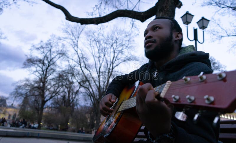 Hombre negro que toca una guitarra y que canta la sentada feliz en un banco de un parque fotografía de archivo libre de regalías Hombre negro que toca una guitarra y que canta la sentada feliz en un banco de un parque fotografía de archivo libre de regalías