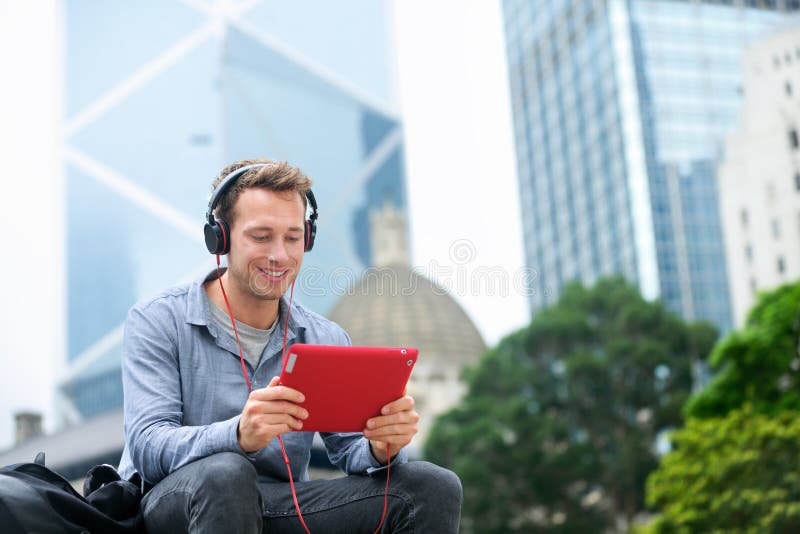 Man talking on tablet pc having video chat conversation in sitting outside using app on 4g wireless device wearing headphones. Casual young urban professional male in his late 20s. Hong Kong. Man talking on tablet pc having video chat conversation in sitting outside using app on 4g wireless device wearing headphones. Casual young urban professional male in his late 20s. Hong Kong.