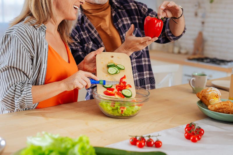 Cutting vegetables. Man holding big red pepper while wife cutting vegetables and putting them into bowl. Cutting vegetables. Man holding big red pepper while wife cutting vegetables and putting them into bowl