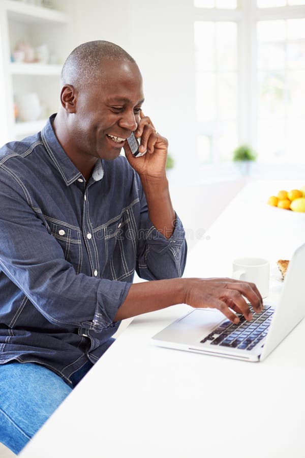 Hombre que usa el ordenador portátil y hablando en el teléfono en cocina en casa imagen de archivo libre de regalías Hombre que usa el ordenador portátil y hablando en el teléfono en cocina en casa imagen de archivo libre de regalías