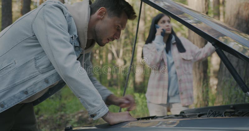 Hombre reparando un auto roto mientras una mujer pide servicio de emergencia pidiendo ayuda en el bosque