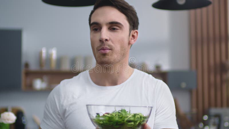 Hombre sonriente comiendo ensalada fresca en la cocina de casa. deportista disfrutando de comida saludable almacen de metraje de vídeo