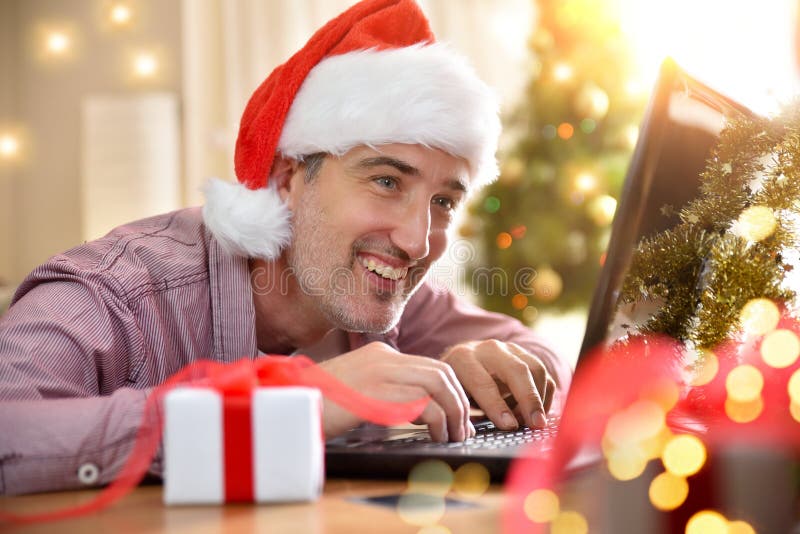 Hombre sonriente con sombrero de navidad usando laptop en casa imagenes de archivo
