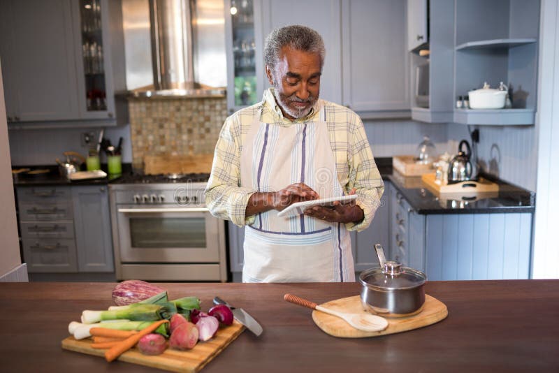 Hombre sonriente que usa la tableta mientras que cocina la comida imagen de archivo Hombre sonriente que usa la tableta mientras que cocina la comida imagen de archivo