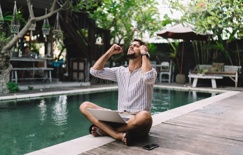 Hombre triste con ordenador portátil apretando los puños mientras está sentado en la piscina imagen de archivo