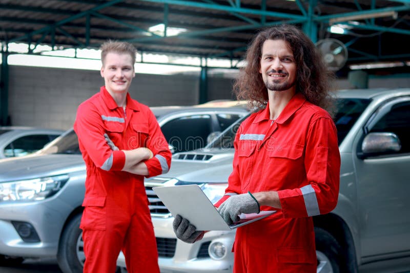 Hombres mecánicos guapos con uniforme rojo que sostienen computadora portátil de pie en el taller mecánico de vehículos que trabaj. Hombres mecánicos guapos con uniforme rojo que sostienen computadora portátil de pie en el taller mecánico de vehículos trabajo en equipo de mecánico de coches que trabajan juntos reparando el automóvil del cliente en la tienda de servicios de automóviles