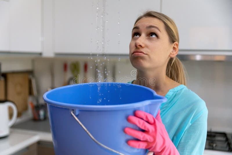 Home flooded by upstairs or roof damage - woman holding bucket while water leaking from ceiling in kitchen