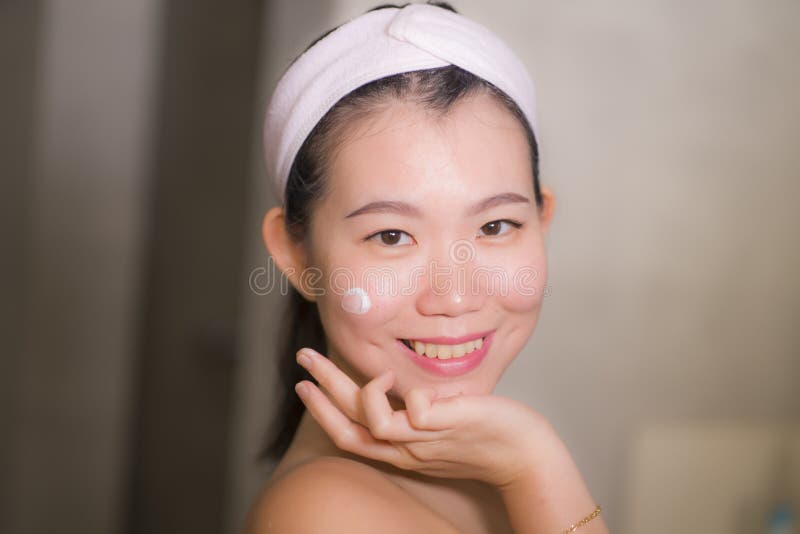 Home lifestyle portrait of young happy and beautiful Asian Korean woman at bathroom mirror applying skin care moisturizer and anti