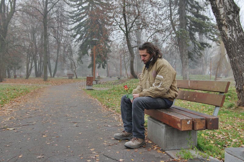 Homeless and hoping for miracle. A thoughtful sad homeless young man sitting on a bench in the park with a beautiful rose in hands, and hoping for a miracle stock photos Homeless and hoping for miracle. A thoughtful sad homeless young man sitting on a bench in the park with a beautiful rose in hands, and hoping for a miracle stock photos