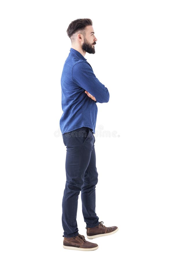 Homem adulto novo na camisa azul da sarja de Nimes que anda e que olha afastado com braços cruzados